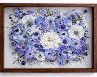 May include: A framed floral arrangement featuring a variety of dried flowers in shades of blue, purple, and white. The flowers are arranged in a dense, layered composition within a dark brown wooden frame. The arrangement includes anemones and other delicate blooms.