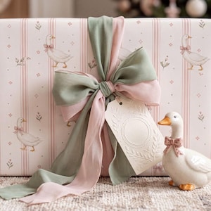 May include: A rectangular gift box wrapped in light pink paper with vertical stripes and a repeating pattern of white geese wearing pink bows. A large sage green and pink fabric bow is tied around the box with a blank, embossed cream gift tag.