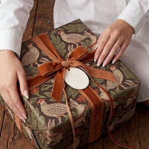 May include: A square gift box wrapped in dark brown paper featuring a repeating pattern of pheasants, rabbits, and green foliage. The box is tied with a wide, copper-coloured velvet ribbon and features a small, oval, off-white embossed gift tag.
