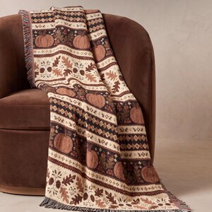 Può includere: Una coperta tessuta con un motivo ripetuto di zucche, foglie di quercia e ghiande nei toni del marrone, crema e arancione scuro. La coperta presenta un bordo sfrangiato e fasce orizzontali di forme geometriche alternate a motivi botanici.