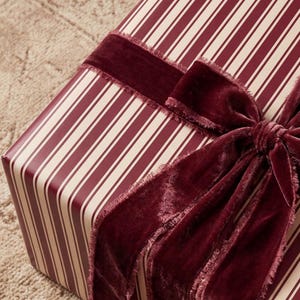 Könnte beinhalten: Eine rechteckige Geschenkbox, eingewickelt in Papier mit einem Muster aus vertikalen burgunderroten und cremefarbenen Streifen. Die Box ist mit einem breiten, tief burgunderroten Samtband umwickelt, das zu einer Schleife mit ausgefransten Kanten gebunden ist.