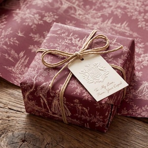 Könnte beinhalten: Eine quadratische Geschenkbox, eingewickelt in dunkelrotes Papier mit weißem Toile-de-Jouy-Muster, gebunden mit beigefarbener Schnur. Ein cremefarbener, rechteckiger Geschenkanhänger mit geprägtem Wappen trägt den Text: To: My Love, From: Your Cupid.