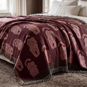 May include: A deep burgundy woven throw blanket featuring a repeating pattern of light pink stylized cat figures. The blanket has a textured appearance with fringed edges along the bottom and sides, draped over a bed with white pillows.