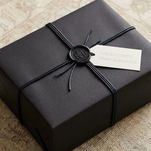 May include: A rectangular gift box wrapped in matte black paper, secured with thin black cord and a circular black wax seal featuring the letters RL. A small white rectangular gift tag is attached, printed with the text FOR: ELIZABETH FROM: ALEXANDER.