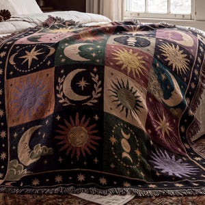 May include: A woven throw blanket featuring a grid pattern of celestial motifs including suns, moons, and stars in shades of black, green, purple, and gold. The blanket has a fringed edge and displays various lunar phases and sun faces.