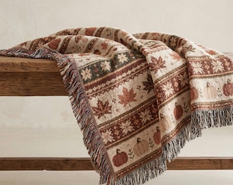 May include: A woven throw blanket featuring a repeating pattern of pumpkins, maple leaves, and floral motifs in shades of brown, cream, and orange. The blanket has a textured finish with decorative fringe along the edges, showcasing a detailed design.