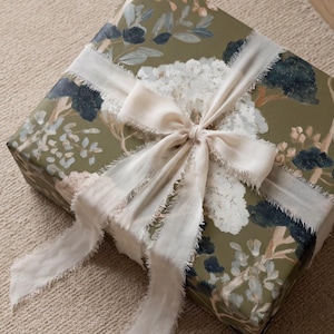 May include: A square gift box wrapped in olive green paper featuring a painted floral pattern of white, dark blue, and light pink blossoms. The box is tied with a wide, off-white silk ribbon that has frayed, raw edges.
