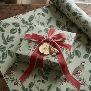 May include: A rectangular gift box wrapped in light green paper featuring a botanical pattern of dark green leaves, brown branches, and red berries. The box is tied with a dusty rose velvet ribbon, a dried pear slice, and a small bird ornament.