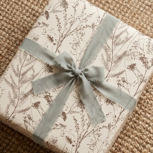 May include: A wrapped gift with a cream-colored botanical print paper and a gray ribbon tied in a bow. The paper features a repeating pattern of brown floral and plant illustrations. The ribbon has a frayed edge.