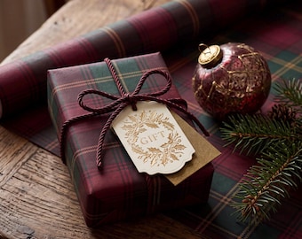 May include: A rectangular gift box wrapped in dark red and green plaid paper, tied with a dark red twisted cord. A cream-colored tag with a gold foil holly wreath design and the word GIFT is attached. A red ornament and pine sprigs are nearby.