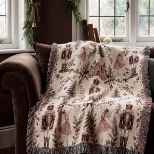 May include: A cream-colored woven throw blanket featuring a repeating pattern of nutcracker figures, ballet dancers in pink dresses, castles, and evergreen trees in shades of brown and muted pink, finished with a decorative fringed edge.