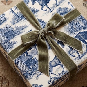 May include: A square gift box wrapped in cream-colored paper featuring a blue toile pattern of horses, riders, and rustic cabins. The package is tied with a wide, olive green velvet ribbon that has frayed edges and is knotted in a bow.