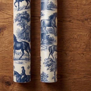 May include: Two rolls of wallpaper featuring a cream background with a repeating blue toile pattern. The design depicts horses, riders, trees, and small buildings in a detailed, monochromatic illustrative style across the cylindrical surfaces.