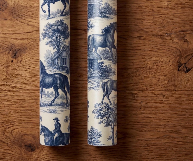 May include: Two rolls of wallpaper featuring a cream background with a repeating blue toile pattern. The design depicts horses, riders, trees, and small buildings in a detailed, monochromatic illustrative style across the cylindrical surfaces.