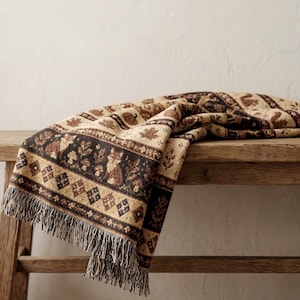 Puede incluir: Una manta tejida con un patrón repetitivo de zorros, ardillas, calabazas y hojas de arce en tonos marrón, beige y crema. La manta tiene un acabado texturizado con un borde decorativo de diamantes y un borde con flecos en la parte inferior.