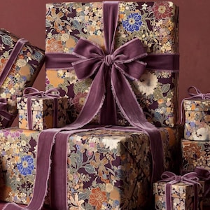 Könnte beinhalten: Eine Sammlung von quadratischen und rechteckigen Geschenkboxen, eingewickelt in dunkelviolettes Papier mit einem dichten Blumenmuster aus blauen, orangefarbenen und goldenen Blüten. Jede Box ist mit einem breiten Samtband in Pflaume gebunden.