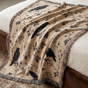 May include: A woven throw blanket in a tan colour featuring a repeating pattern of black crows perched on dark floral branches. The blanket has a textured fabric construction with frayed edges along the sides and a detailed woven design throughout.