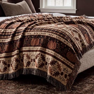 May include: A woven throw blanket featuring a repeating pattern of pumpkins, oak leaves, and acorns in shades of brown, tan, and cream. The blanket has a textured surface with horizontal bands of geometric designs and a fringed edge along the bottom.