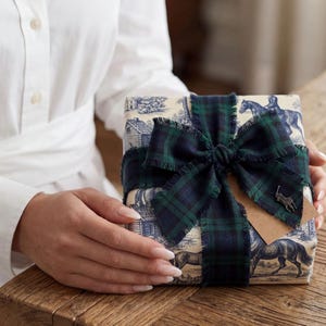 May include: A square gift box wrapped in cream-colored paper featuring a blue toile de jouy pattern of horses and houses. The box is tied with a dark green and navy blue plaid ribbon with frayed edges and a small metal horse logo charm attached.