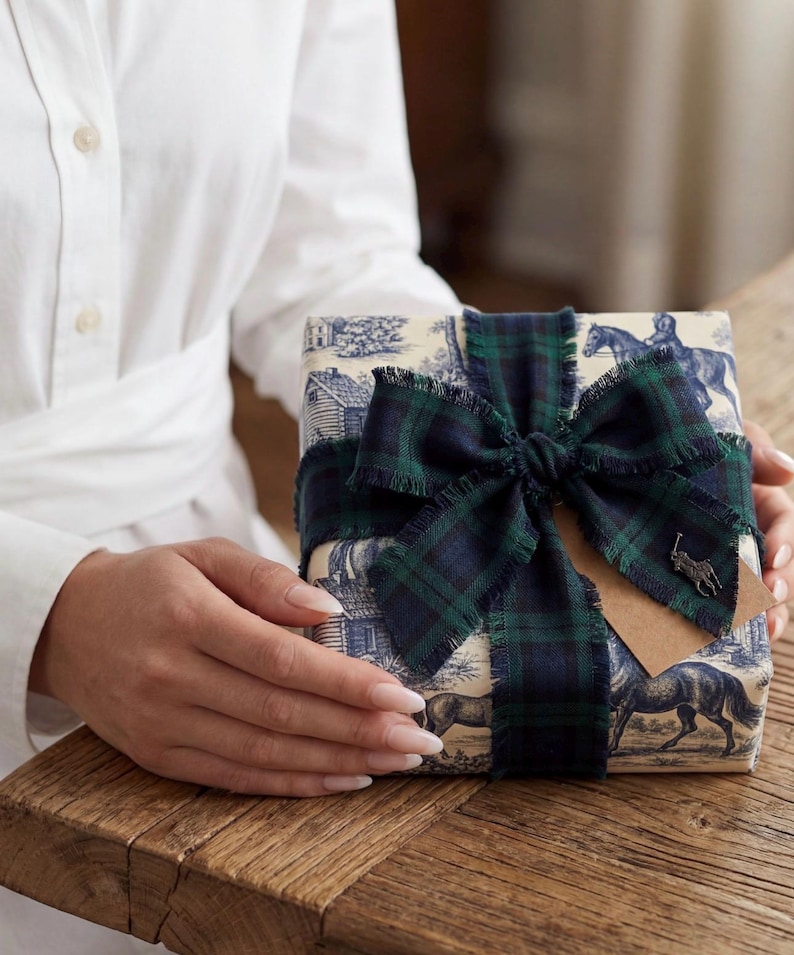 May include: A square gift box wrapped in cream-colored paper featuring a blue toile de jouy pattern of horses and houses. The box is tied with a dark green and navy blue plaid ribbon with frayed edges and a small metal horse logo charm attached.
