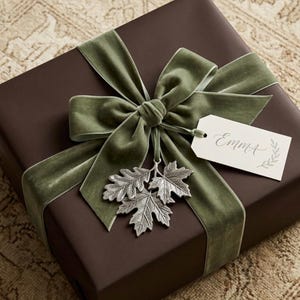 May include: A square gift box wrapped in dark brown paper, tied with a wide sage green velvet ribbon. A metallic silver ornament shaped like three oak leaves hangs from the center knot, alongside a white rectangular gift tag inscribed with Emma.
