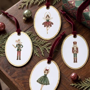 May include: Four oval-shaped hanging ornaments with gold glitter edges and dark red velvet ribbons. Each ornament features a white center with a watercolor illustration: a mouse soldier, a fairy dancer, a nutcracker, and a ballerina in a green dress.