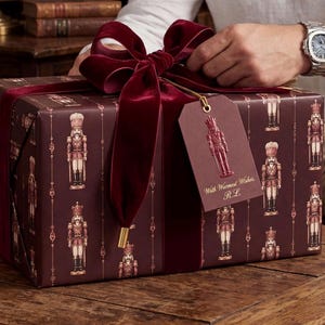 May include: A rectangular gift box wrapped in dark burgundy paper featuring a repeating nutcracker pattern, tied with a wide dark red velvet ribbon. A matching burgundy gift tag hangs from the ribbon, printed with a nutcracker and the text With Warmest Wishes, R.L.