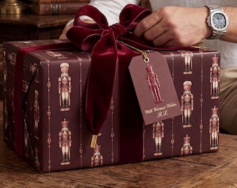 May include: A rectangular gift box wrapped in dark burgundy paper featuring a repeating nutcracker pattern, tied with a wide dark red velvet ribbon. A matching burgundy gift tag hangs from the ribbon, printed with a nutcracker and the text With Warmest Wishes, R.L.