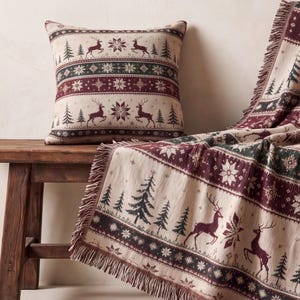 May include: A square throw pillow and a matching woven throw blanket featuring a repeating pattern of dark red deer, dark green pine trees, and white snowflakes on a cream-colored background, with fringed edges on the throw blanket.