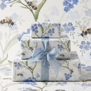 Pode incluir: Três caixas de presente empilhadas, embrulhadas em papel branco com um desenho floral e de abelhas. As caixas são amarradas com uma fita azul clara. O fundo apresenta um padrão floral correspondente com abelhas.