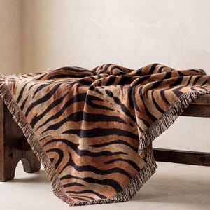 Puede incluir: Una manta rectangular tejida con un patrón repetitivo de rayas de tigre en tonos negro, marrón oscuro y beige. La manta tiene una superficie texturizada y está terminada con detalles de flecos cortos deshilachados en los cuatro bordes.