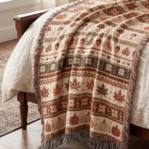 Könnte beinhalten: Eine gewebte Wohndecke mit einem sich wiederholenden Muster aus Kürbissen, Pilzen, Eicheln, Ahornblättern und Blumenmotiven in Orange-, Braun- und Cremetönen. Die Decke hat eine strukturierte Oberfläche und einen Fransenrand an den Seiten und unten.