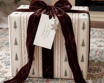 May include: A square gift box wrapped in cream-colored paper with a pattern of green pine trees and thin red stripes. A large, dark brown velvet bow sits on top, accompanied by a white rectangular gift tag with script text and a snowflake charm.