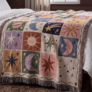 May include: A woven throw blanket featuring a grid of square panels with celestial motifs including smiling suns, crescent moons, stars, planets, and constellations in shades of pink, blue, green, yellow, and purple with fringed edges.