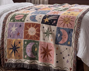 May include: A woven throw blanket featuring a grid of square panels with celestial motifs including smiling suns, crescent moons, stars, planets, and constellations in shades of pink, blue, green, yellow, and purple with fringed edges.