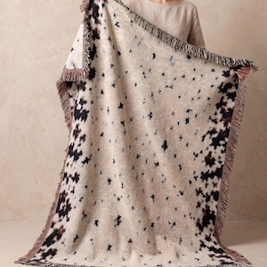 May include: A woven throw blanket featuring a cream-coloured base with a scattered black and dark brown abstract spotted pattern. The rectangular blanket is finished with a decorative fringe along all four edges, creating a textured appearance.