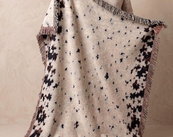 May include: A woven throw blanket featuring a cream-colored base with a scattered black and dark brown abstract spotted pattern. The rectangular blanket is finished with a decorative fringe along all four edges, creating a textured appearance.