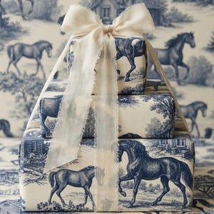 May include: A stack of three rectangular gift boxes wrapped in cream-colored paper featuring a blue toile de jouy pattern of horses and rural landscapes. The stack is tied with a wide, frayed-edge cream ribbon tied in a bow at the top.