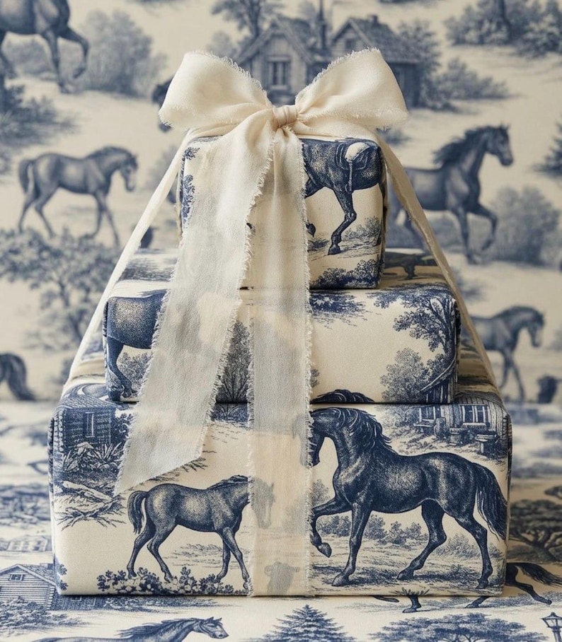 May include: A stack of three rectangular gift boxes wrapped in cream-colored paper featuring a blue toile de jouy pattern of horses and rural landscapes. The stack is tied with a wide, frayed-edge cream ribbon tied in a bow at the top.