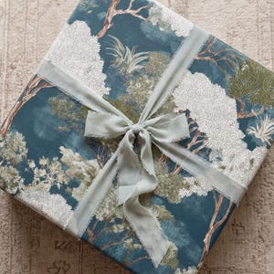 May include: A square gift box wrapped in dark teal paper featuring a detailed white and green botanical forest print. The package is tied with a light sage green frayed-edge ribbon that is knotted into a bow at the center of the top surface.