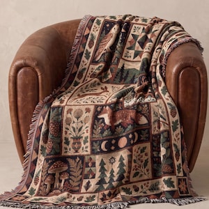 Puede incluir: Una manta tejida con un diseño de patchwork de ilustraciones con temática forestal que incluye un búho, un zorro, un conejo, setas, pinos, piñas y fases lunares en tonos tierra de marrón, verde, crema y azul oscuro con flecos.