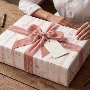 May include: A rectangular gift box wrapped in white paper featuring a pattern of pink stripes and illustrations of ducks wearing bows. The box is tied with a wide, dusty pink velvet ribbon and includes a cream-colored tag labeled Ralph Lauren.