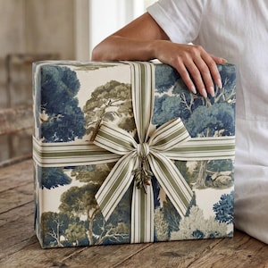 May include: A rectangular gift box wrapped in cream-colored paper featuring a blue and green forest landscape print. The box is tied with a striped olive green and white ribbon, finished with a decorative metal olive branch charm at the center.