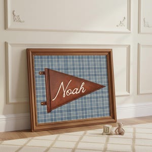 May include: A framed wall art piece featuring a brown leather pennant with the name "Noah" in white script. The pennant is set against a blue and white plaid background within a wooden frame. The artwork is leaning against a white wall.
