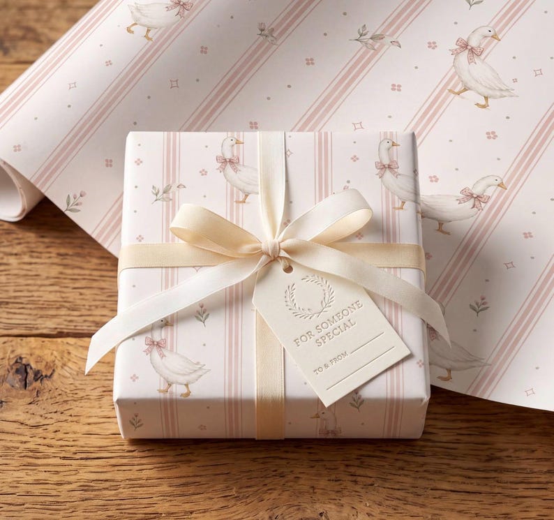 May include: A rectangular gift box wrapped in light pink and white striped paper featuring illustrations of white geese wearing pink bows. The box is tied with a cream-colored satin ribbon and includes a gift tag printed with FOR SOMEONE SPECIAL.