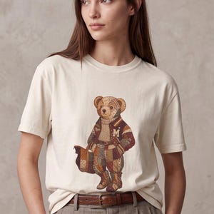 May include: A cream-coloured short-sleeve t-shirt featuring a graphic print of a teddy bear dressed in a patchwork varsity jacket, a turtleneck sweater, and boots. The bear holds a brown bag and has the letter M on the jacket chest.