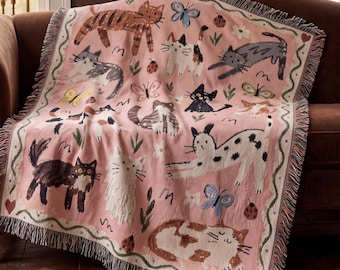 May include: A woven throw blanket in a soft pink hue featuring a repeating pattern of various cats in shades of brown, gray, black, and white, interspersed with colorful butterflies, small ladybugs, and white flowers, finished with a fringed edge.