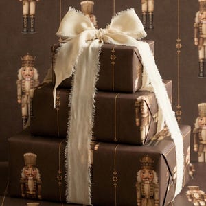May include: A stack of three rectangular gift boxes wrapped in dark brown paper featuring a repeating nutcracker pattern with vertical gold lines. The boxes are tied together with a wide, cream-colored ribbon with frayed edges.