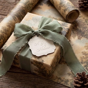May include: A square gift box wrapped in beige paper with a brown landscape print, tied with a sage green frayed silk ribbon. A white scalloped gift tag is attached to the ribbon, featuring the embossed text A Gift of Nature in script.