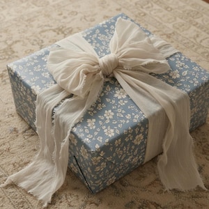 May include: A square gift box wrapped in blue paper featuring a white floral pattern. The box is tied with a large, off-white fabric ribbon that has frayed, raw edges. The ribbon is knotted in the center, with long ends draping over the sides.